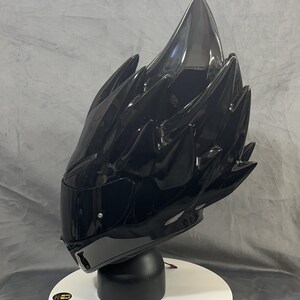 Op de afbeelding: Een glanzend zwarte motorhelm met een uniek, stekelig ontwerp. De helm staat op een witte, cirkelvormige standaard met een zwarte basis. Het vizier is donker en de algehele uitstraling is futuristisch en edgy.