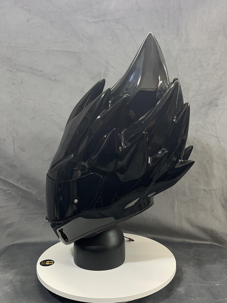 Op de afbeelding: Een glanzend zwarte motorhelm met een uniek, stekelig ontwerp. De helm staat op een witte, cirkelvormige standaard met een zwarte basis. Het vizier is donker en de algehele uitstraling is futuristisch en edgy.