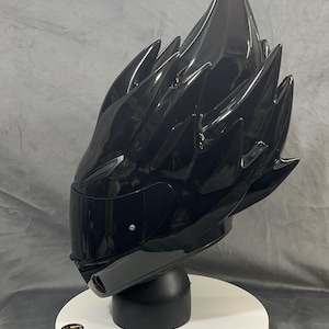 Op de afbeelding: Een glanzend zwarte motorhelm met een uniek, stekelig ontwerp dat lijkt op gestileerd haar. De helm heeft een getint vizier en wordt gepresenteerd op een zwarte standaard met een witte basis. De algehele esthetiek is futuristisch en edgy.