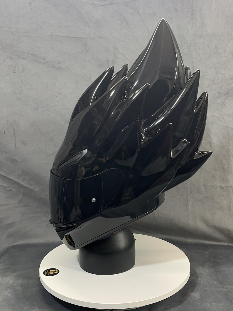 Op de afbeelding: Een glanzend zwarte motorhelm met een uniek, stekelig ontwerp dat lijkt op gestileerd haar. De helm heeft een getint vizier en wordt gepresenteerd op een zwarte standaard met een witte basis. De algehele esthetiek is futuristisch en edgy.