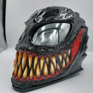 May include: Black motorcycle helmet with a red and yellow Venom design. The helmet has a large, toothy grin and a silver visor.