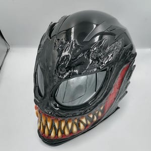 May include: Black motorcycle helmet with a detailed Venom design. The helmet has a red and black paint scheme with a large, toothy grin.