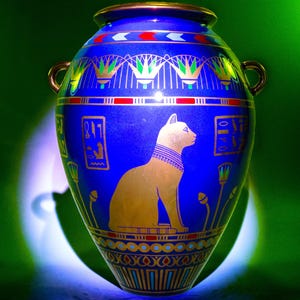 May include: A blue ceramic vase with gold accents and an Egyptian-style design featuring a cat. The vase has a wide mouth and two handles.