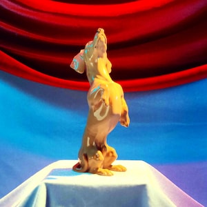 May include: A glossy, light brown ceramic figurine of a dachshund standing on its hind legs with its head tilted upward. The dog has long, floppy ears and a smooth, reflective surface that catches the light, showcasing its elongated body shape.