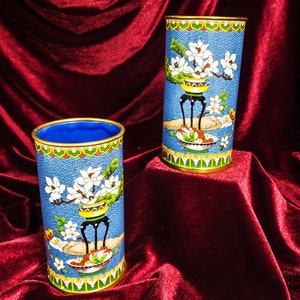 May include: Two blue and white cloisonné vases with floral designs. The vases are cylindrical and have a gold rim. They are sitting on a red velvet surface.