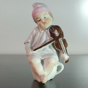 May include: A porcelain figurine of a child wearing a pink hat and playing a violin. The child is sitting on a white cup.