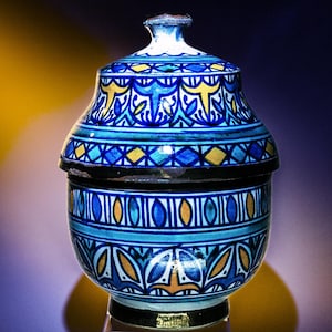 Peut inclure: Un pot en céramique bleu et blanc avec un couvercle, décoré de motifs géométriques et floraux complexes en jaune et bleu. Le pot a un corps rond et un petit couvercle rond.