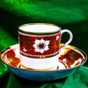 Op de afbeelding: Een wit theekopje en schotel met een rood en gouden bloemenpatroon. Het kopje heeft een gouden rand en een wit interieur. De schotel heeft een bijpassend patroon en een blauwe rand.