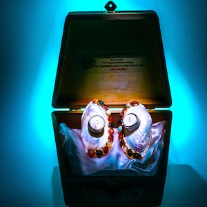 May include: A wooden jewelry box with a hinged lid, open to reveal two large, iridescent pearl shell earrings. Each earring is adorned with colorful gemstones and a central button. The box sits on a blue surface.