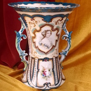 May include: A large, white porcelain vase with ornate gold and blue painted designs. The vase has two handles and features a portrait of a woman in a white dress.