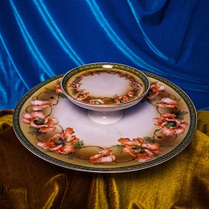 May include: A two-tiered serving dish with a white background and a floral design featuring red poppies. The dish is made of porcelain and has a green and gold trim.