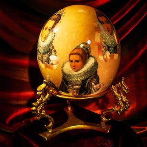 May include: A decorative egg with portraits of women in period clothing, set on a gold-colored stand. The egg is a light yellow color with the portraits in shades of brown, white, and black. The stand has ornate, scroll-like details.