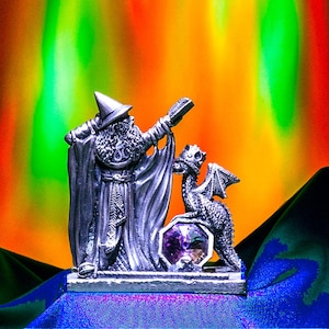 May include: A pewter figurine of a wizard holding a staff with a dragon beside him. The wizard is wearing a pointed hat and a long robe. The dragon is standing on a purple gem. The figurine is set against a colorful background.
