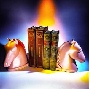 Puede incluir: Dos sujetalibros de cabeza de caballo rosa sosteniendo un conjunto de cinco libros. Los libros están encuadernados en cuero marrón con letras doradas. Los libros de la izquierda y la derecha están titulados "Mark Twain Autobiography" y los tres libros del medio están titulados "Adventures of Huckleberry Finn".
