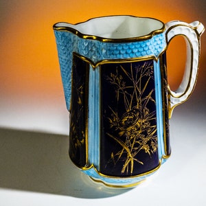 Puede incluir: Una jarra de porcelana azul y dorada con un diseño floral. La jarra tiene un borde festoneado y un asa.