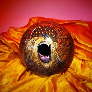 Peut inclure: Une gourde ronde et brune ornée d'une illustration peinte à la main représentant un ours rugissant, gueule ouverte et dents visibles, sur un fond sombre à motifs avec des accents circulaires dorés, posée sur un tissu orange drapé.