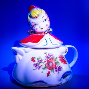 以下が含まれることがあります： 花柄の白い陶器製のティーポット。赤い帽子をかぶった赤と白の陶器製のドールが蓋になっています。ドールは黄色い髪をしています。