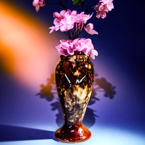 以下が含まれることがあります： まだら模様の釉薬がかかった茶色と白の陶器の花瓶に、ピンクの花が活けられている