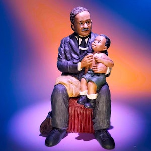 May include: A figurine of a man in a suit holding a young child. The man is sitting on a red stool and the child is sitting on his lap. The figurine is made of painted resin and has a realistic appearance.