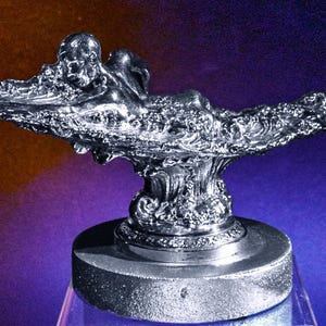 May include: A silver-colored car hood ornament featuring a stylized figure reclining on a wave-like base.