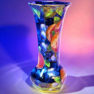 May include: A tall, flared ceramic vase featuring a deep blue background decorated with vibrant, hand-painted illustrations of various fruits, including peaches and grapes, accented with green leaves and a gold-toned rim at the top opening.