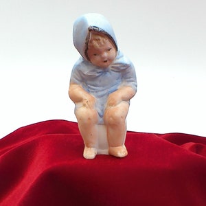 May include: A small porcelain figurine of a child wearing a blue bonnet and dress, sitting on a white toilet.