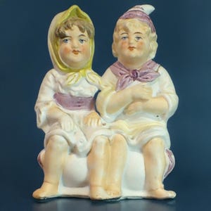 May include: A porcelain figurine of two children sitting on a white base. The girl is wearing a yellow and white dress with a purple sash and a yellow bonnet. The boy is wearing a white shirt with a purple sash and a purple and white hat.