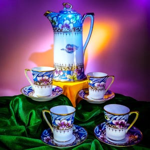 Könnte beinhalten: Ein Porzellan-Teeservice mit einer hohen, blau geblümten Teekanne und vier passenden Tassen und Untertassen. Die Tassen und die Teekanne haben goldfarbene Verzierungen. Das Set ist auf einem grünen Samttuch mit lila und gelbem Hintergrund angeordnet.