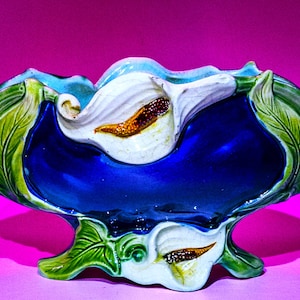 May include: A ceramic centerpiece bowl with a vibrant design. The bowl features a deep blue interior, white calla lily accents, and green leaves. The base is supported by a green, leaf-shaped pedestal.