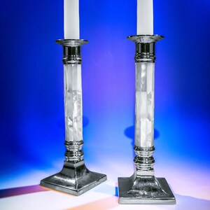 Pode incluir: Dois castiçais de prata com velas brancas. Os castiçais têm uma base branca texturizada e um topo prateado.