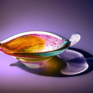 May include: A colorful glass bowl with a unique shape and a handle on one side. The bowl has swirls of orange, yellow, green, and pink glass.