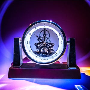 Puede incluir: Un reloj Bulova plateado con una esfera transparente y engranajes visibles. El reloj está sobre una base de madera oscura y está iluminado por una luz morada y naranja.