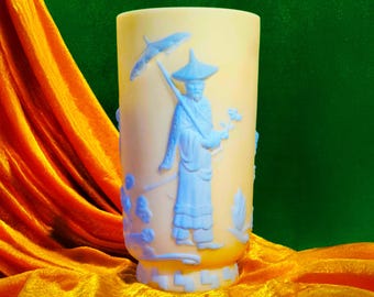 May include: A cylindrical, pale yellow glass vase with a light blue relief design of an Asian figure holding an umbrella and flowers. The vase has a flared base and is set against a backdrop of orange and green fabric.