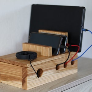 Puede incluir: Estación de carga de madera con ranuras para varios dispositivos, incluyendo un portátil, teléfonos y un reloj inteligente. La estación tiene un sistema de gestión de cables integrado con agujeros para los cables.