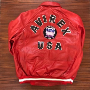 Può includere: Giubbotto bomber in pelle rossa con una grande toppa "AVIREX ALL AMERICANS USA" sul retro. La toppa presenta uno scudo con una bandiera americana e l'anno 1975.