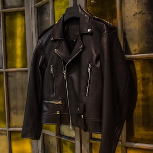 Puede incluir: Chaqueta de cuero negra de estilo motero clásico. La chaqueta presenta un cuello con muescas, cierre de cremallera asimétrico, múltiples bolsillos con cremallera y charreteras. Cuelga de una percha negra.