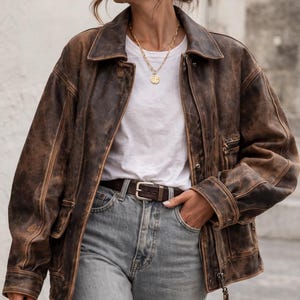 Op de afbeelding: Een bruine, verweerde leren jas met ritssluiting, gedragen over een wit t-shirt en een lichtblauwe jeans. Een gouden ketting en bruine riem maken de look compleet. De jas heeft een klassieke kraag en meerdere zakken.