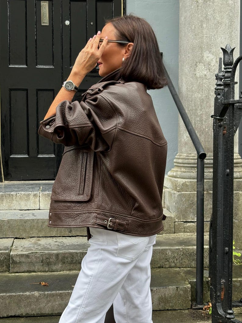 Side view of a woman wearing a vintage brown oversized leather jacket made from genuine sheepskin leather, featuring a relaxed fit, drop shoulders, textured pebbled leather finish, utility flap pocket, adjustable waist buckle, and retro-inspired silhouette. Styled with white jeans for a chic streetwear look, this women's oversized leather jacket showcases timeless 90s fashion, vintage biker jacket details, premium leather craftsmanship, casual luxury outerwear, fall winter fashion, handmade leat