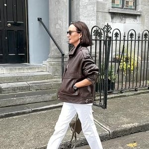 Side view of a woman wearing a vintage brown oversized leather jacket made from genuine sheepskin leather, featuring a relaxed fit, drop shoulders, textured pebbled leather, utility pockets, adjustable waist tabs, and retro-inspired details. Styled with white jeans and minimalist sandals, this women's oversized leather jacket captures 90s streetwear fashion, vintage biker jacket style, casual luxury outerwear, brown leather coat, handmade leather jacket, retro fashion aesthetic, premium leather