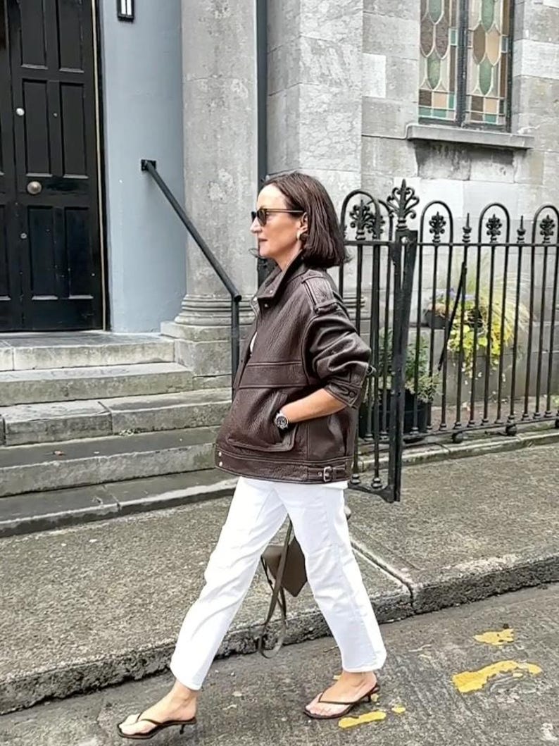 Side view of a woman wearing a vintage brown oversized leather jacket made from genuine sheepskin leather, featuring a relaxed fit, drop shoulders, textured pebbled leather, utility pockets, adjustable waist tabs, and retro-inspired details. Styled with white jeans and minimalist sandals, this women's oversized leather jacket captures 90s streetwear fashion, vintage biker jacket style, casual luxury outerwear, brown leather coat, handmade leather jacket, retro fashion aesthetic, premium leather