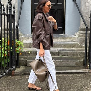 Woman wearing a vintage brown oversized leather jacket crafted from genuine sheepskin leather, styled with white straight-leg jeans, minimalist heels, and a taupe leather shoulder bag. Features a relaxed oversized fit, drop shoulders, textured pebbled leather, utility flap pockets, adjustable waist tabs, and retro-inspired tailoring. This women's brown leather jacket embodies 90s fashion, vintage streetwear, oversized biker jacket style, casual luxury outerwear, handmade leather jacket, premium