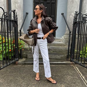 Front view of a woman wearing a vintage brown oversized leather jacket crafted from genuine sheepskin leather, featuring a relaxed fit, drop shoulders, structured collar, textured pebbled leather finish, utility pockets, and retro-inspired detailing. Styled with white jeans and a minimalist leather tote bag, this women's leather jacket embodies 90s fashion, vintage streetwear, oversized biker jacket style, casual luxury outerwear, timeless brown leather coat, handmade leather jacket, fall winter