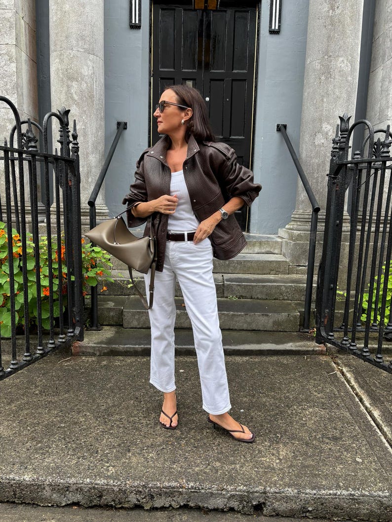 Front view of a woman wearing a vintage brown oversized leather jacket crafted from genuine sheepskin leather, featuring a relaxed fit, drop shoulders, structured collar, textured pebbled leather finish, utility pockets, and retro-inspired detailing. Styled with white jeans and a minimalist leather tote bag, this women's leather jacket embodies 90s fashion, vintage streetwear, oversized biker jacket style, casual luxury outerwear, timeless brown leather coat, handmade leather jacket, fall winter