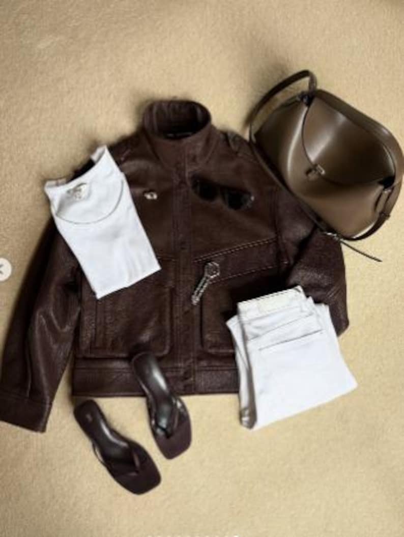Flat lay outfit featuring a women's vintage brown oversized leather jacket crafted from genuine sheepskin leather, styled with a white t-shirt, white straight-leg jeans, minimalist sandals, sunglasses, and a taupe leather shoulder bag. This retro-inspired leather jacket showcases a relaxed oversized fit, textured pebbled leather finish, utility pockets, and timeless vintage detailing. Perfect for 90s fashion lovers, vintage streetwear enthusiasts, casual luxury outfits, oversized biker jacket