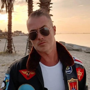 May include: A man wearing a black leather jacket with a brown fur collar and multiple patches. The jacket has a white t-shirt underneath. The man is wearing sunglasses and is standing in front of a beach with palm trees in the background.