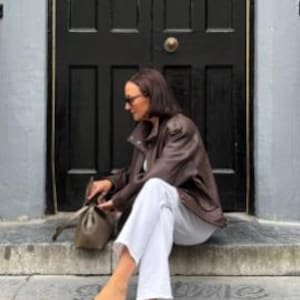 Woman seated on stone steps wearing a vintage brown oversized leather jacket made from genuine sheepskin leather, featuring a relaxed fit, drop shoulders, textured pebbled leather finish, utility pockets, and retro-inspired detailing. Styled with white straight-leg jeans, minimalist sandals, and a taupe leather shoulder bag, this women's leather jacket showcases 90s fashion, vintage streetwear style, oversized biker jacket aesthetic, casual luxury outerwear, handmade leather jacket, premium brow