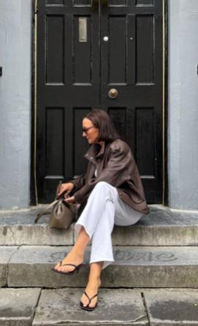 Woman seated on stone steps wearing a vintage brown oversized leather jacket made from genuine sheepskin leather, featuring a relaxed fit, drop shoulders, textured pebbled leather finish, utility pockets, and retro-inspired detailing. Styled with white straight-leg jeans, minimalist sandals, and a taupe leather shoulder bag, this women's leather jacket showcases 90s fashion, vintage streetwear style, oversized biker jacket aesthetic, casual luxury outerwear, handmade leather jacket, premium brow