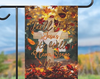 Puede incluir: Una bandera de jardín con el texto "Fall For Jesus He Will Never Leave You" sobre un fondo de hojas de otoño y una cruz de madera. La bandera se muestra en un soporte de metal negro, con un cielo azul y follaje de otoño en el fondo.