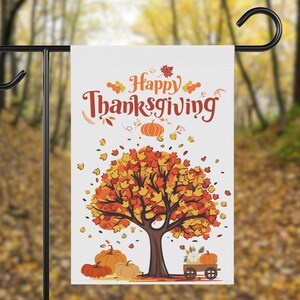 以下が含まれることがあります： オレンジと赤のスクリプトで「Happy Thanksgiving」という言葉が書かれた白いガーデンフラッグ。オレンジと黄色の葉、カボチャ、カボチャと花でいっぱいのワゴンが描かれています。黒い金属製のスタンドに吊るされています。