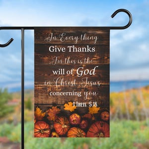 May include: A garden flag with a wooden plank design, featuring the text "In Everything Give Thanks". The flag has a fall theme with pumpkins and leaves. It is displayed on a black metal stand, perfect for outdoor decoration.